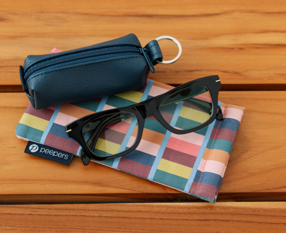 Black glasses on a colorful cloth with a blue case and 'peepers' label on a wooden surface