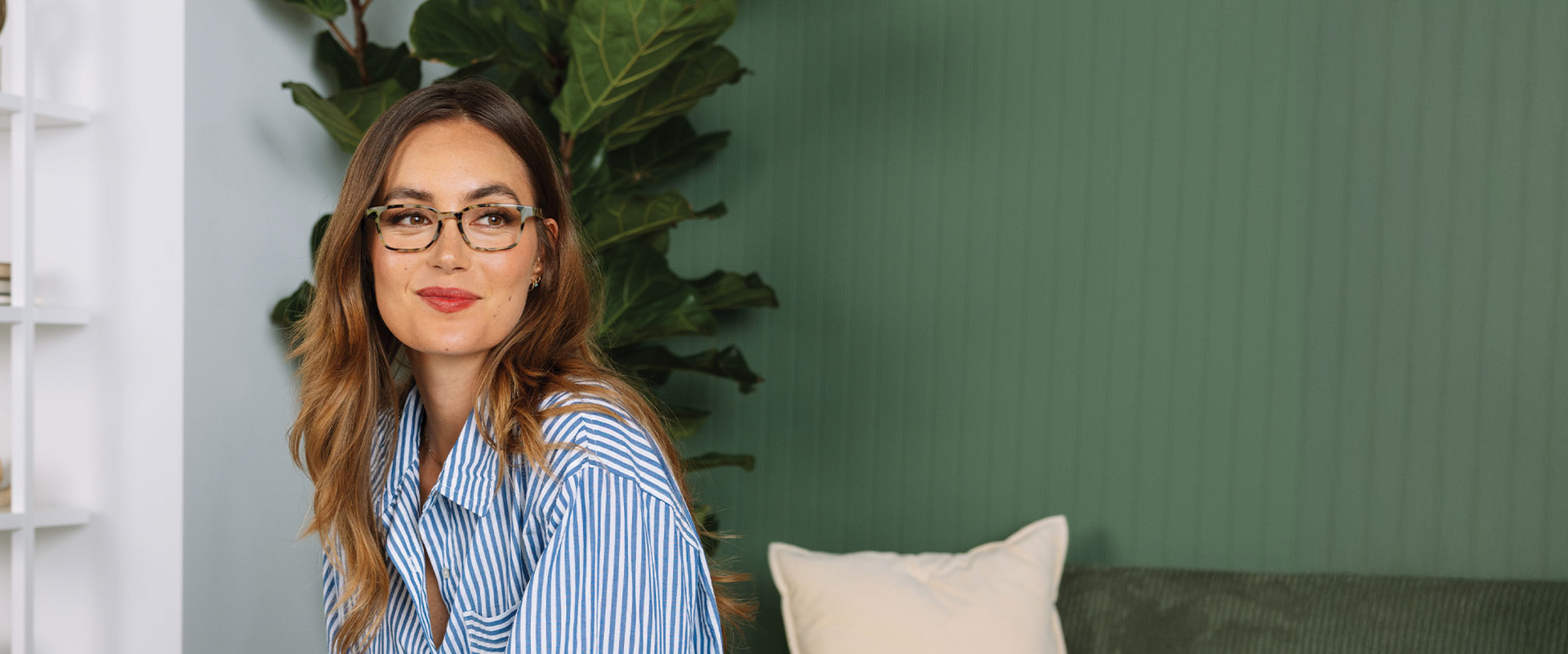 Woman wearing glasses and a striped shirt in a room with green walls and a plant.