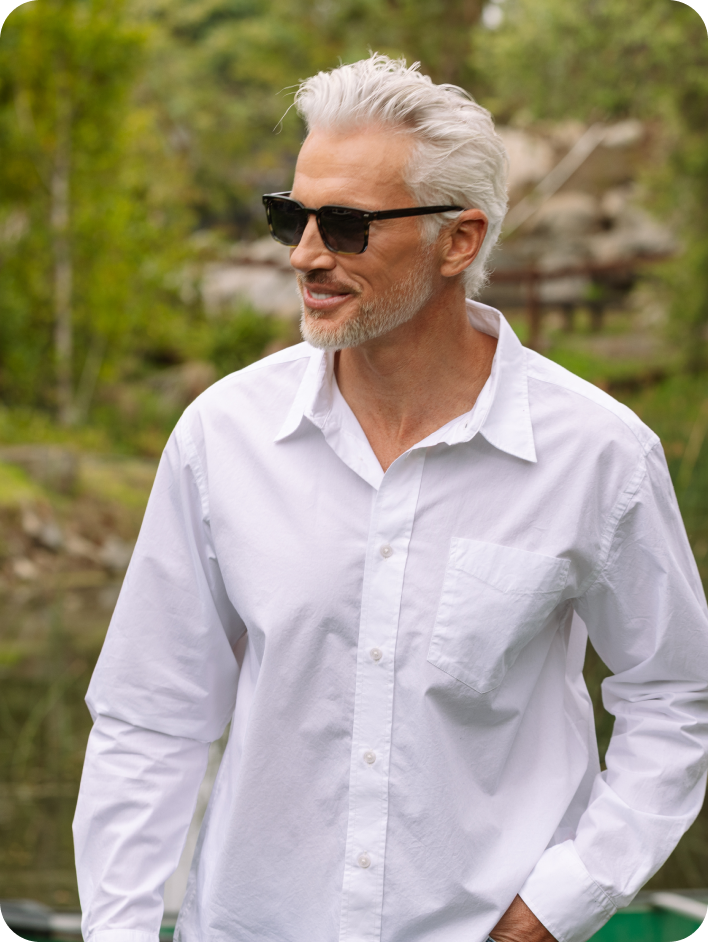 Man wearing a white shirt and sunglasses by a body of water with trees in the background