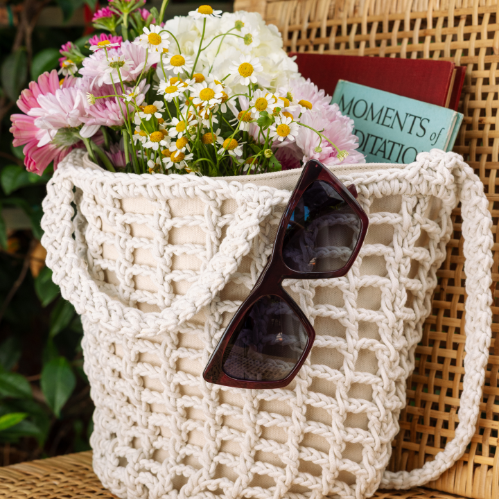 White woven bag with flowers, sunglasses, and a book on a wicker chair.