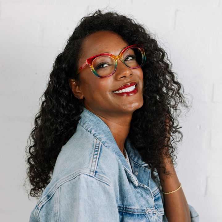 Woman wearing colorful glasses and a denim jacket against a white background