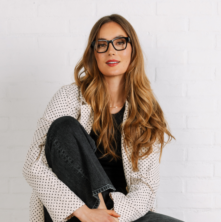 Woman wearing glasses and a polka dot jacket against a white brick wall