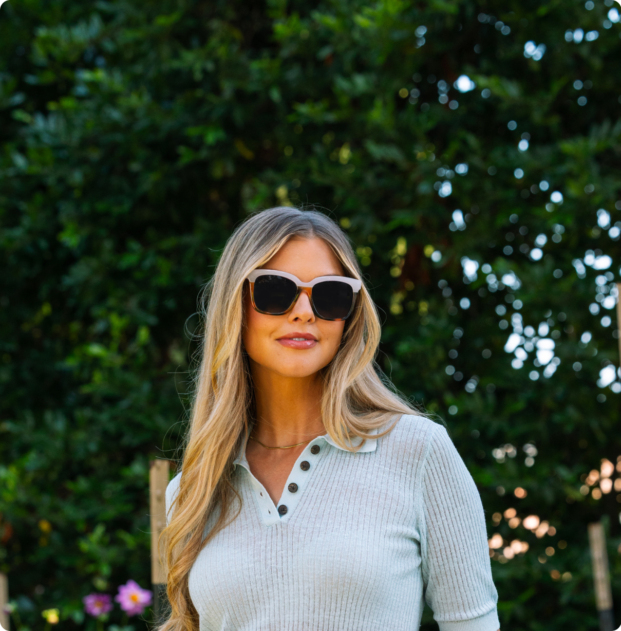 Woman wearing sunglasses and a light-colored shirt with a green, leafy background