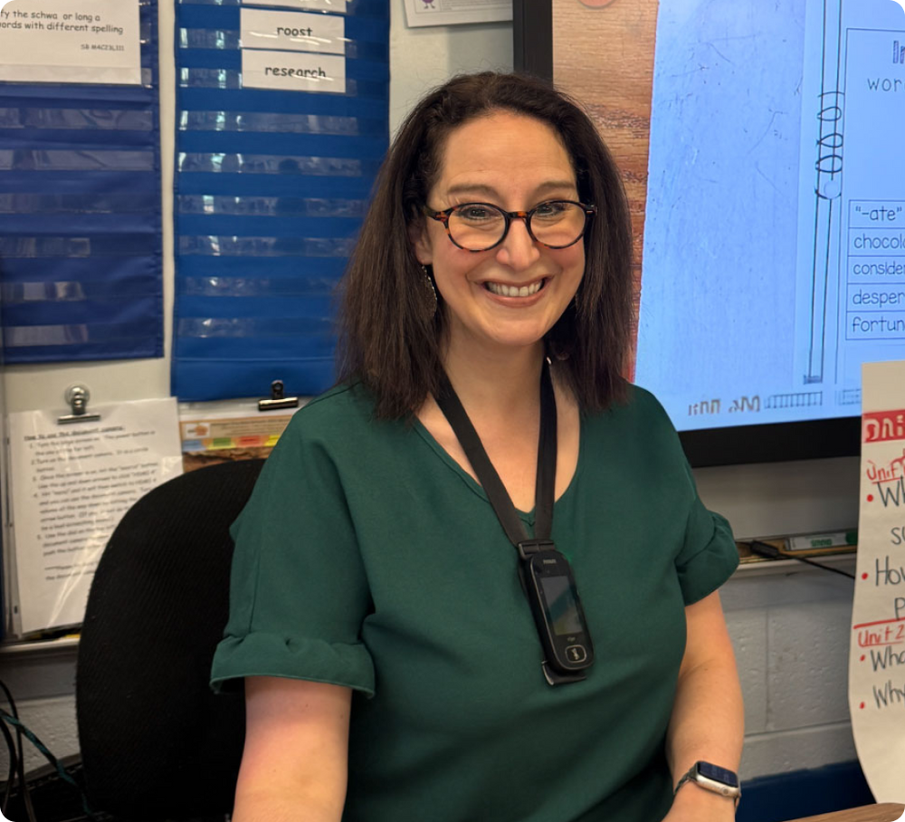 A teacher smiling in her classroom with Peepers eyewear, bulletin board behind her