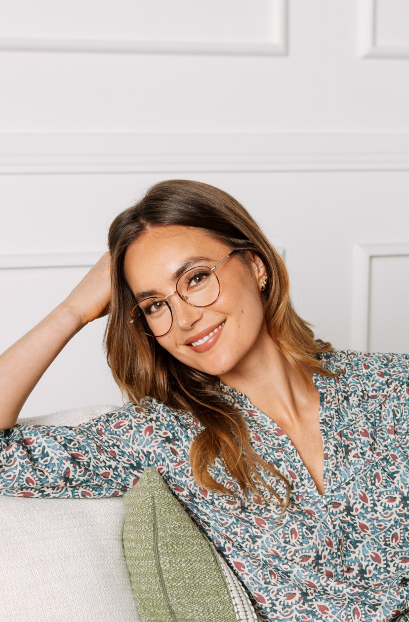 Woman wearing glasses and a floral shirt sitting on a couch with a white wall background