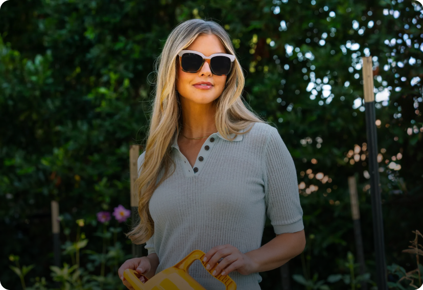 Woman wearing sunglasses and a light-colored shirt outdoors with greenery in the background
